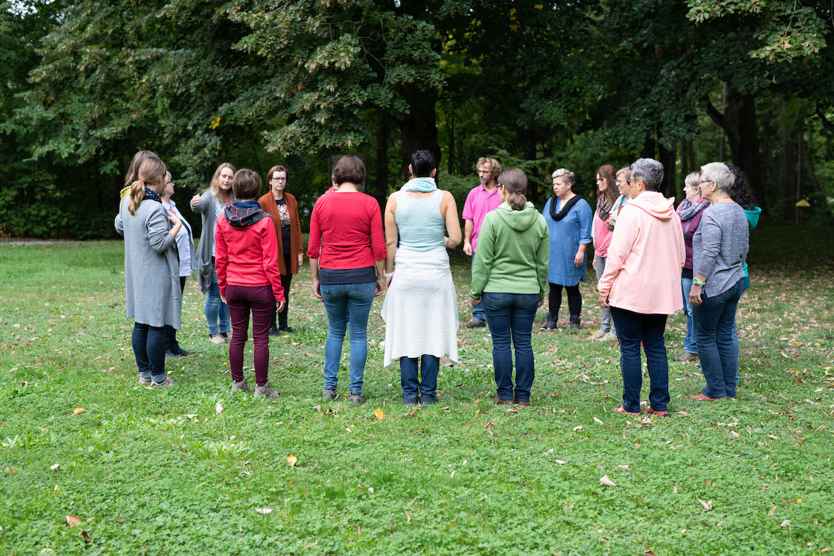 Mehrere Menschen stehen im Kreis. Man sieht vorallem die Rücken. Die Menschen beraten sich. Sie stehen auf einer Wiese, dahinter sieht man einen Wald.